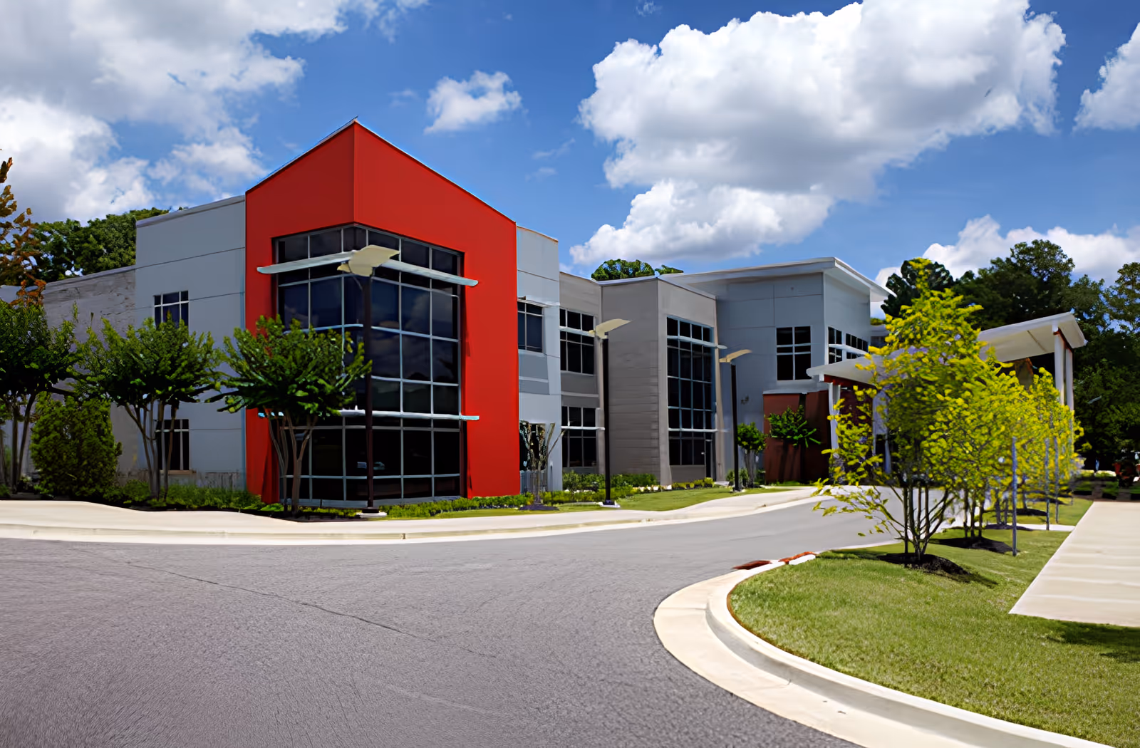 Exterior view of a modern building with a red accent wall and large windows, surrounded by trees and a curved driveway under a partly cloudy blue sky.