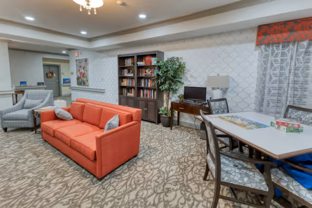 A cozy common area in a senior living facility featuring a red sofa, a gray armchair, a bookshelf filled with books, a potted plant, a small table with a computer monitor and lamp, and a table with chairs set up for games or activities. The room has patterned carpet, wallpaper with a subtle design, and recessed lighting in the ceiling.