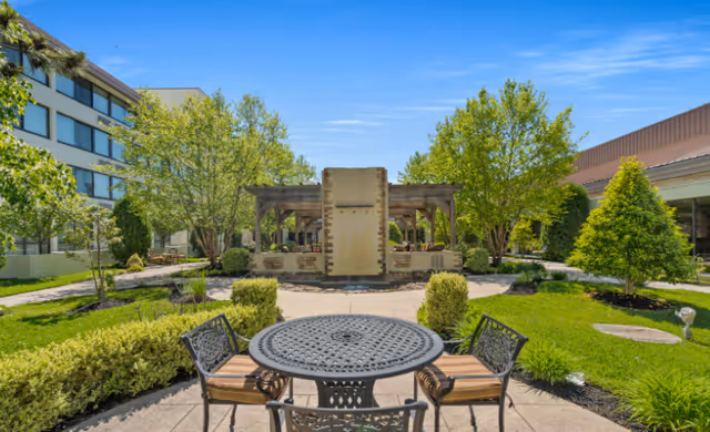 Outdoor patio area at The Addison of Cherry Hill featuring a round metal table with four chairs on a paved surface, surrounded by green grass, bushes, and trees under a clear blue sky. In the background, there is a pergola structure and parts of the building are visible.