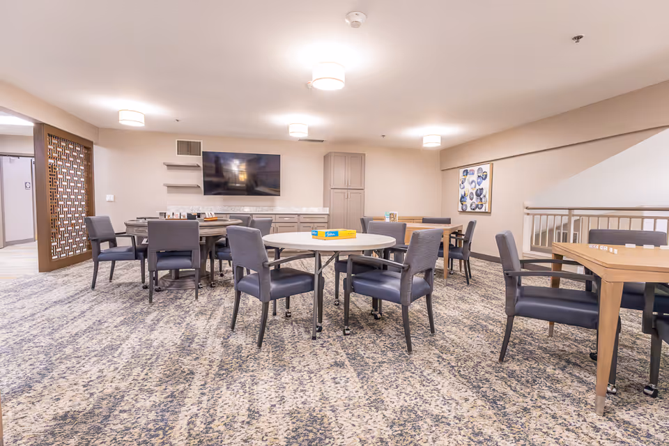 A spacious common room with multiple tables and chairs arranged for group activities or dining. The room features a patterned carpet, neutral-colored walls, a flat-screen TV mounted on the wall, and modern ceiling lights. There is a decorative wooden partition on the left and a framed abstract artwork on the right wall.
