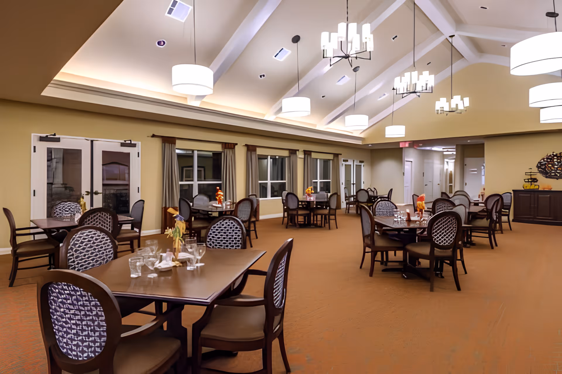 Spacious senior living dining room with multiple tables and chairs under hanging chandeliers and a vaulted ceiling.