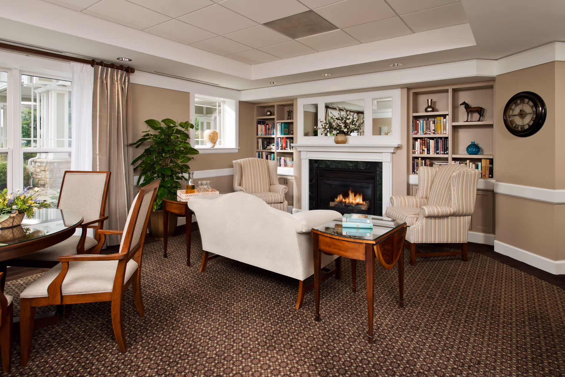 A cozy living room area in a senior living facility featuring a lit fireplace with a white mantel, two striped armchairs, a white upholstered sofa, a wooden side table with books, a round glass-top dining table with wooden chairs, a large green plant, built-in bookshelves with various books and decorative items, a wall clock, and large windows with beige curtains allowing natural light to enter.