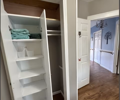 Open white closet with shelves holding folded towels and storage bins, next to an open door labeled with the number 5, leading to a hallway with wooden flooring, white wainscoting, and a decorative wall piece.