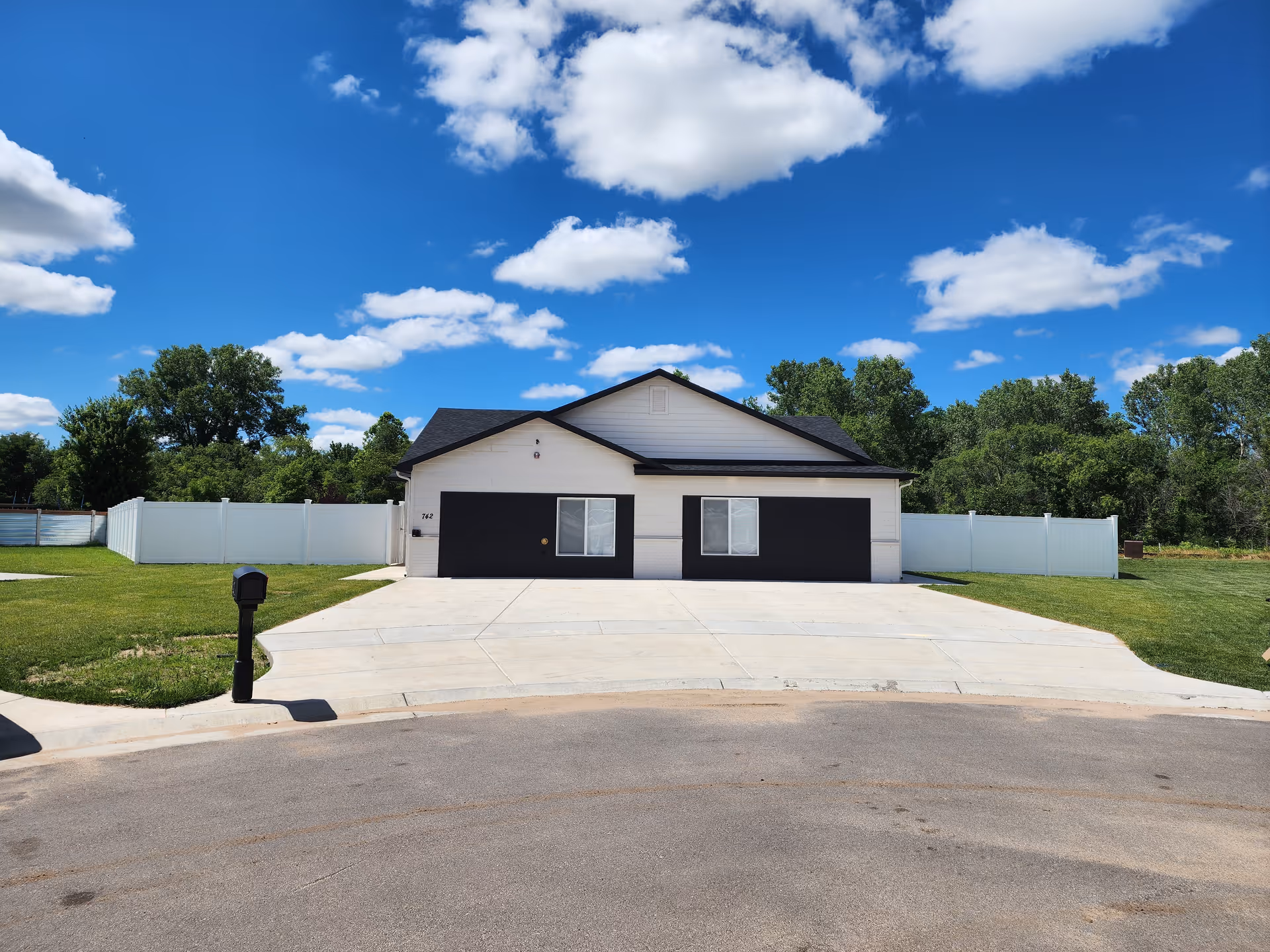 Front exterior of a single-story house with two black garage doors, a wide concrete driveway, white vinyl fence, mailbox and lawn under a blue sky with clouds.