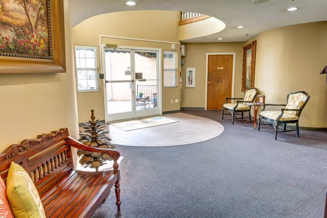 A bright and welcoming lobby area with a wooden bench featuring a yellow cushion on the left, two cushioned chairs with a small table between them on the right, a large mirror on the wall, and glass double doors leading outside. The floor is carpeted with a circular rug near the entrance, and a decorative plant is placed near the bench.