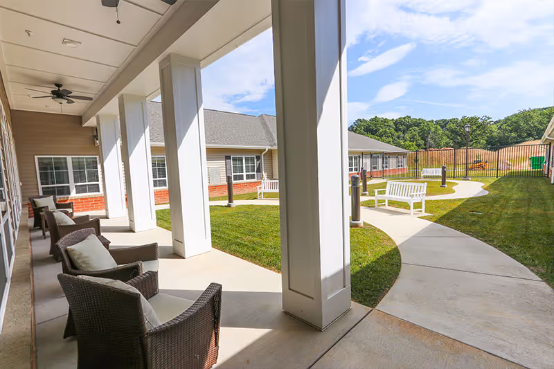 Outdoor patio area at a senior living facility with wicker chairs and cushions arranged along a covered walkway. The patio overlooks a grassy courtyard with a curved concrete pathway, white benches, and a black metal fence enclosing the area. Trees and a partly cloudy sky are visible in the background.