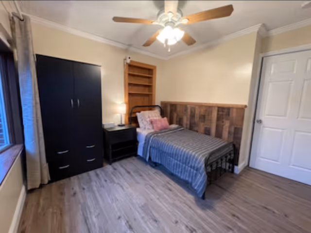 A neatly arranged bedroom with a single bed featuring a gray striped blanket and pink pillows. The bed has a wooden headboard with a rustic design. Next to the bed is a black nightstand with a lamp and a phone. There is a black wardrobe with drawers on the left side of the room. The room has light-colored walls, a ceiling fan with lights, and a window with beige curtains letting in natural light. The floor is covered with wood-like laminate.