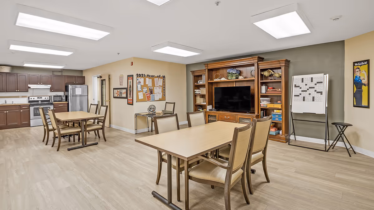 A spacious activity room featuring a dining table with chairs, a kitchenette area with appliances, and a television unit. The walls are decorated with posters and a bulletin board.