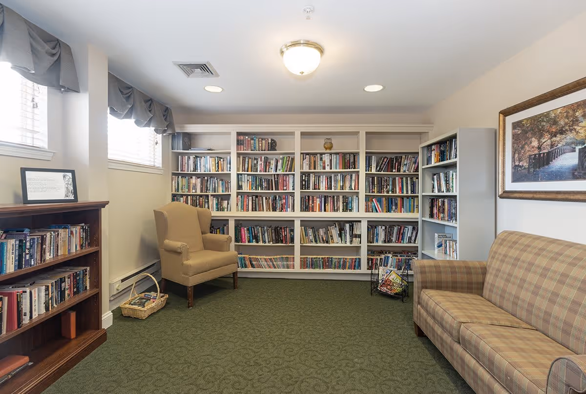 A cozy reading room with green carpet, a beige armchair, a plaid sofa, and multiple bookshelves filled with books. There are two small windows with gray valances, a framed picture on the wall, and a ceiling light fixture.
