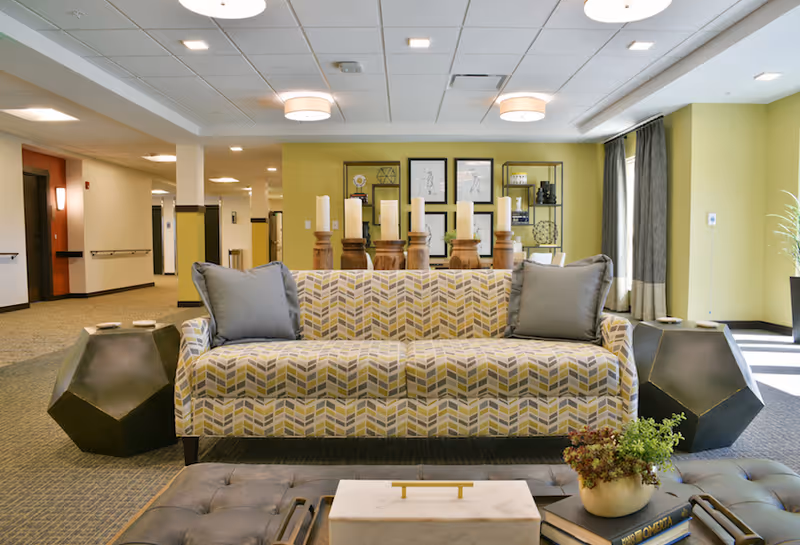 Bright senior living lounge with a patterned sofa flanked by geometric side tables, decorative candles and wall shelving.