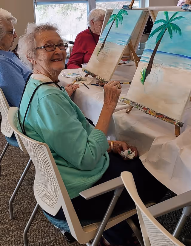 Three elderly women seated at a table in a well-lit room, engaged in painting beach scenes with palm trees on canvases set on easels. One woman in a green sweater is smiling at the camera while holding a paintbrush and a cloth.
