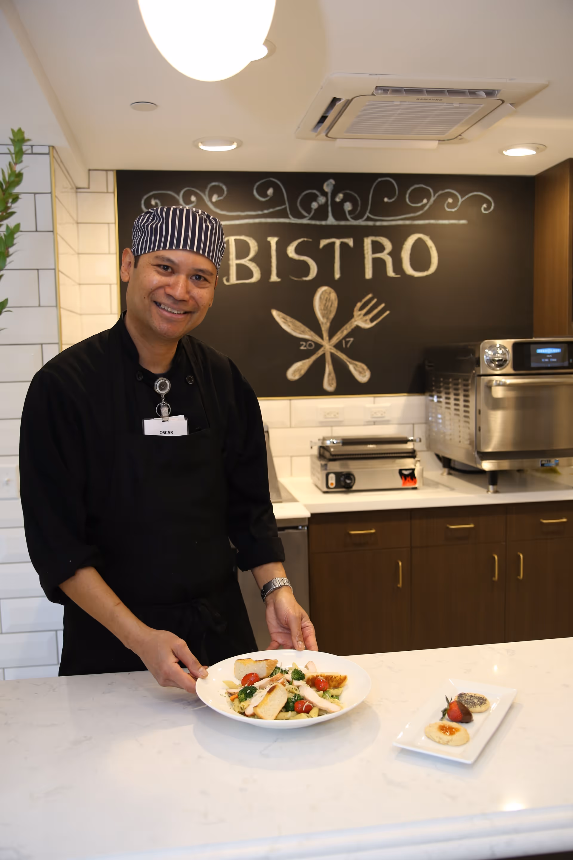 A chef wearing a striped hat and black apron stands behind a kitchen counter holding a plate of salad with bread, cherry tomatoes, and greens. Behind him is a blackboard with the word 'BISTRO' and drawings of a spoon, fork, and knife. The kitchen area has white tiled walls and wooden cabinets with kitchen appliances on the counter.