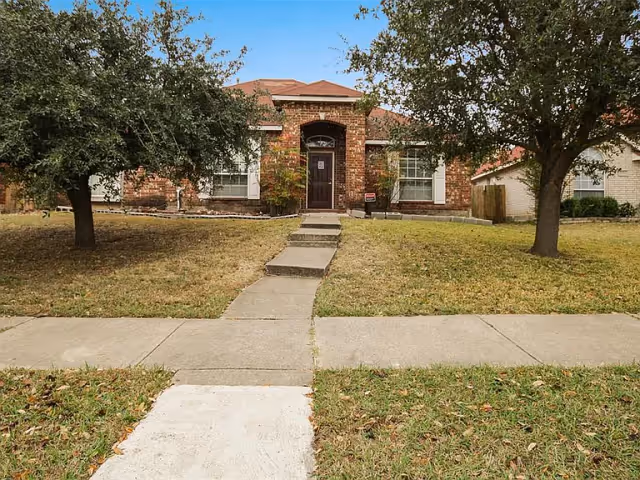 Front exterior view of a single-story brick house with a brown front door, two large windows with white shutters, a concrete pathway leading to the entrance, and two trees on either side of the lawn.