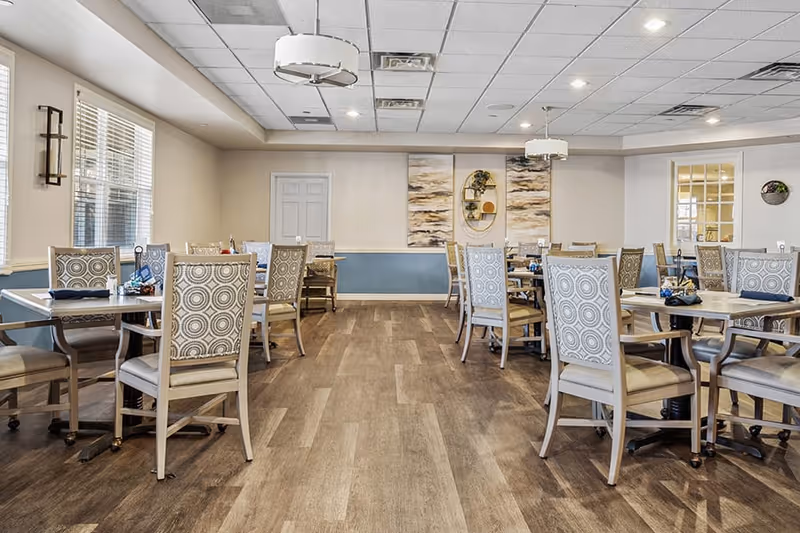 A spacious dining room with multiple tables and chairs arranged neatly. The chairs have patterned backs and cushioned seats. The room features wood flooring, light-colored walls with blue wainscoting, and large windows letting in natural light. Decorative wall art and modern light fixtures hang from the ceiling.
