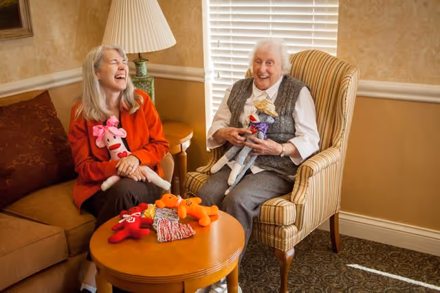 Two elderly women sitting in a cozy living room area, smiling and holding colorful stuffed animals. One woman is seated on a striped armchair, and the other is on a brown sofa. A wooden coffee table with more stuffed animals and a knitted item is in front of them. A lamp and window blinds are visible in the background.