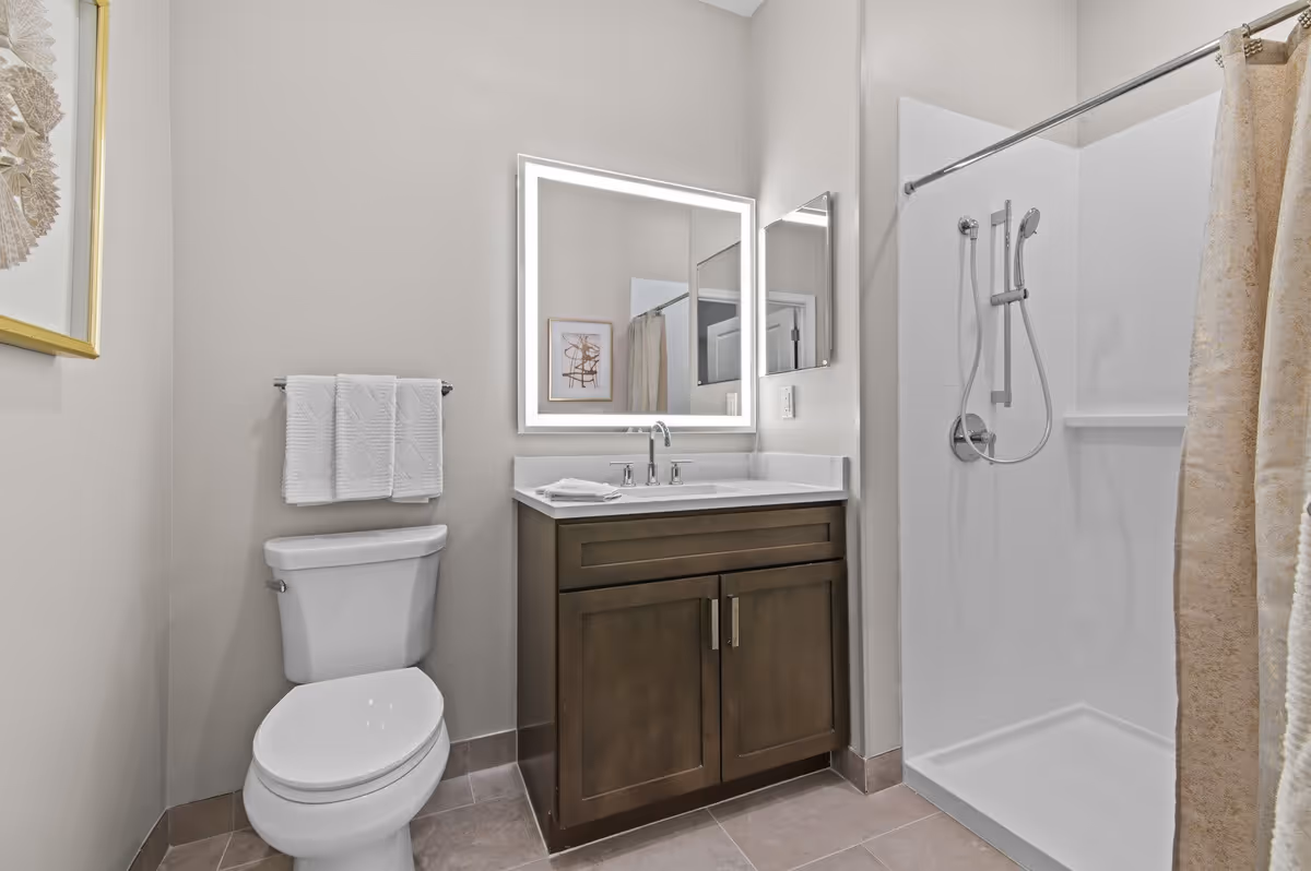 A clean and modern bathroom featuring a white toilet with a towel rack above it holding three white towels, a dark wood vanity with a white countertop and a sink, a large square mirror with built-in lighting above the sink, and a walk-in shower with a handheld showerhead and a beige shower curtain.