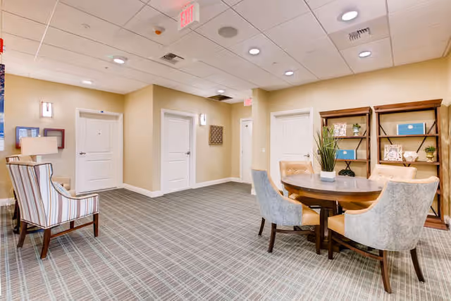 Carpeted communal lounge with a round table and upholstered chairs, shelving, and additional seating near several doors.