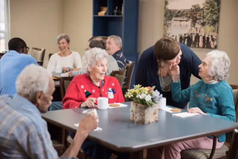 A group of elderly people sitting around a table in a senior living facility dining room, enjoying snacks and drinks while engaging in conversation. A caregiver is interacting warmly with one of the elderly women. The room has a cozy atmosphere with a flower centerpiece on the table and a large photo on the wall in the background.
