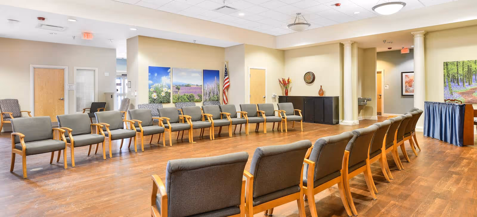 A spacious room with two rows of wooden chairs with gray cushions arranged facing each other on a wooden floor. The room has beige walls, several doors, and decorative wall art including nature-themed paintings. There is an American flag in the corner and a small table with a blue cloth on the right side.