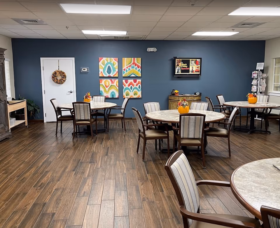 Communal dining/activity room with round tables and chairs, colorful wall art, a wall-mounted TV, and small pumpkin centerpieces.