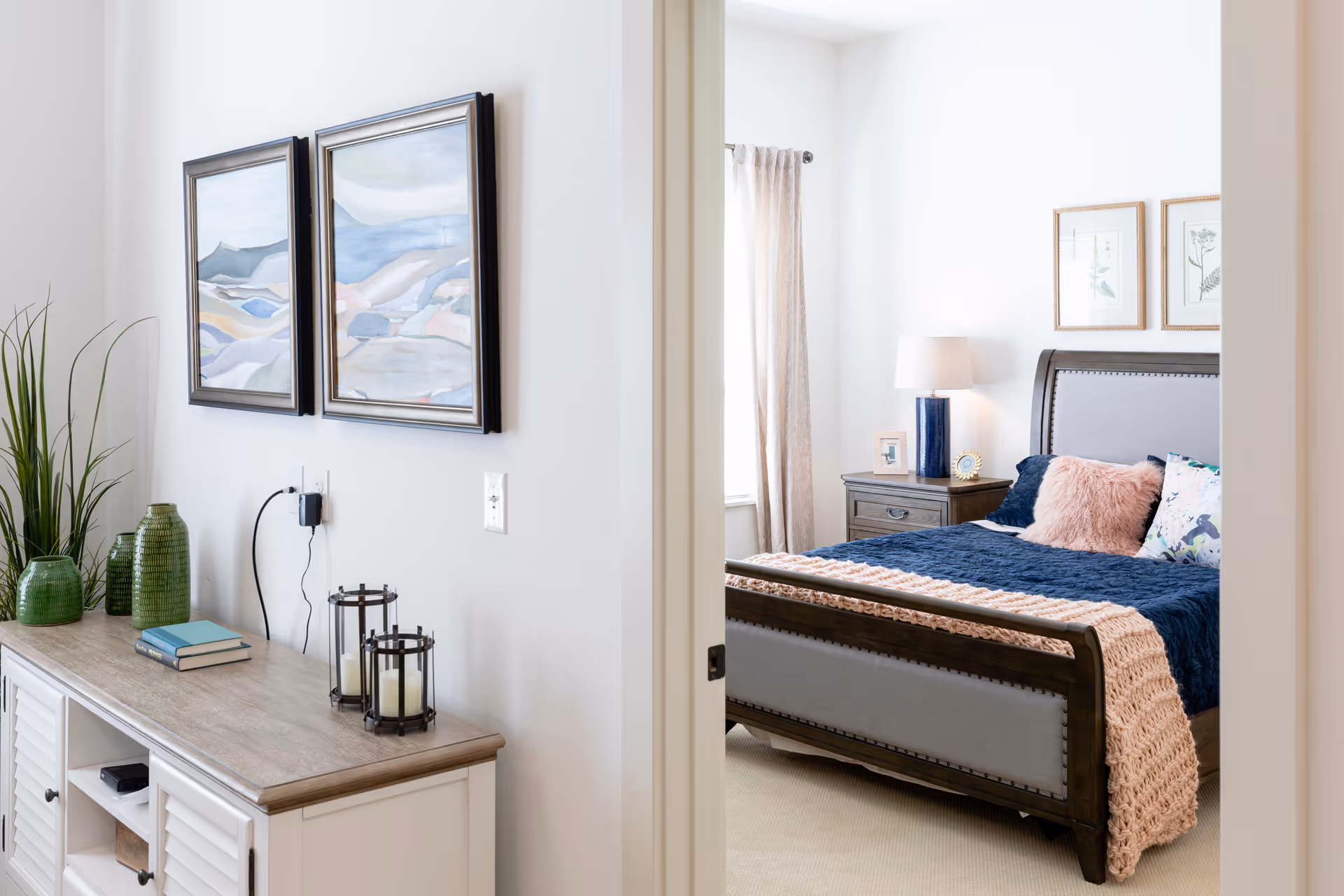 View through an open doorway into a bedroom with a bed featuring a blue blanket and pink throw, a nightstand with a lamp, and framed botanical prints on the wall. Outside the bedroom, there is a console table with green vases, books, and candle holders, along with two abstract landscape paintings on the wall.
