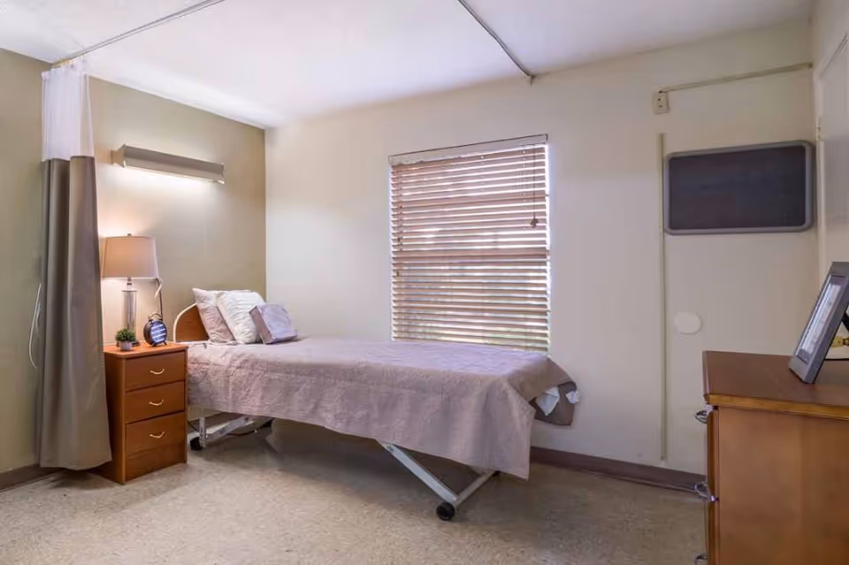 A simple nursing bedroom with a single bed, nightstand and lamp, window blinds and a dresser.