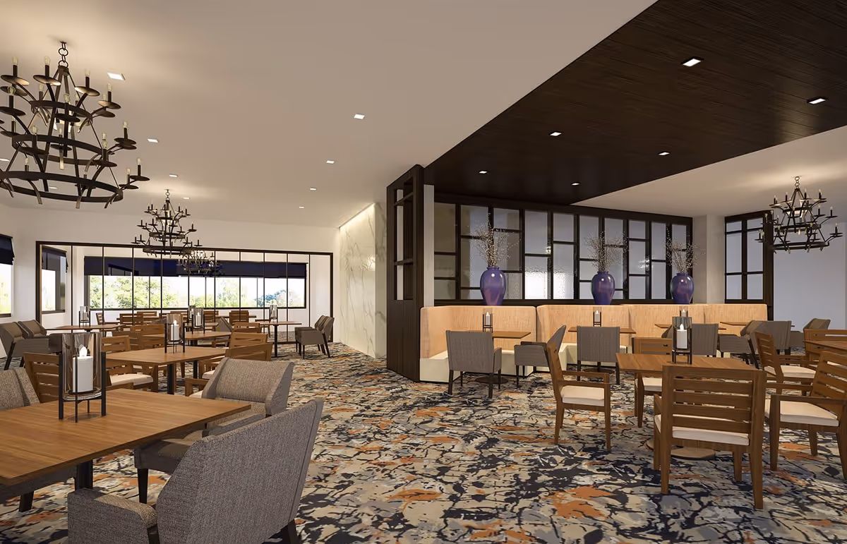 A spacious dining room with multiple wooden tables and chairs arranged neatly. The room features patterned carpet flooring, large windows allowing natural light, and decorative chandeliers hanging from the ceiling. There are also cushioned booth seats along one wall with large blue vases placed on a divider.