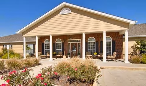 Front entrance of a single-story senior living building with a gabled covered porch, white columns, seating, and landscaped flower beds.