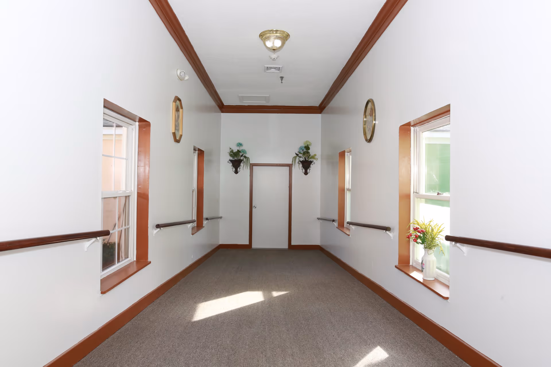 A narrow hallway with white walls and brown trim, featuring handrails on both sides below windows. There are two decorative wall sconces with flowers on the far wall flanking a closed door. The ceiling has a light fixture and ventilation panels, and the floor is carpeted.