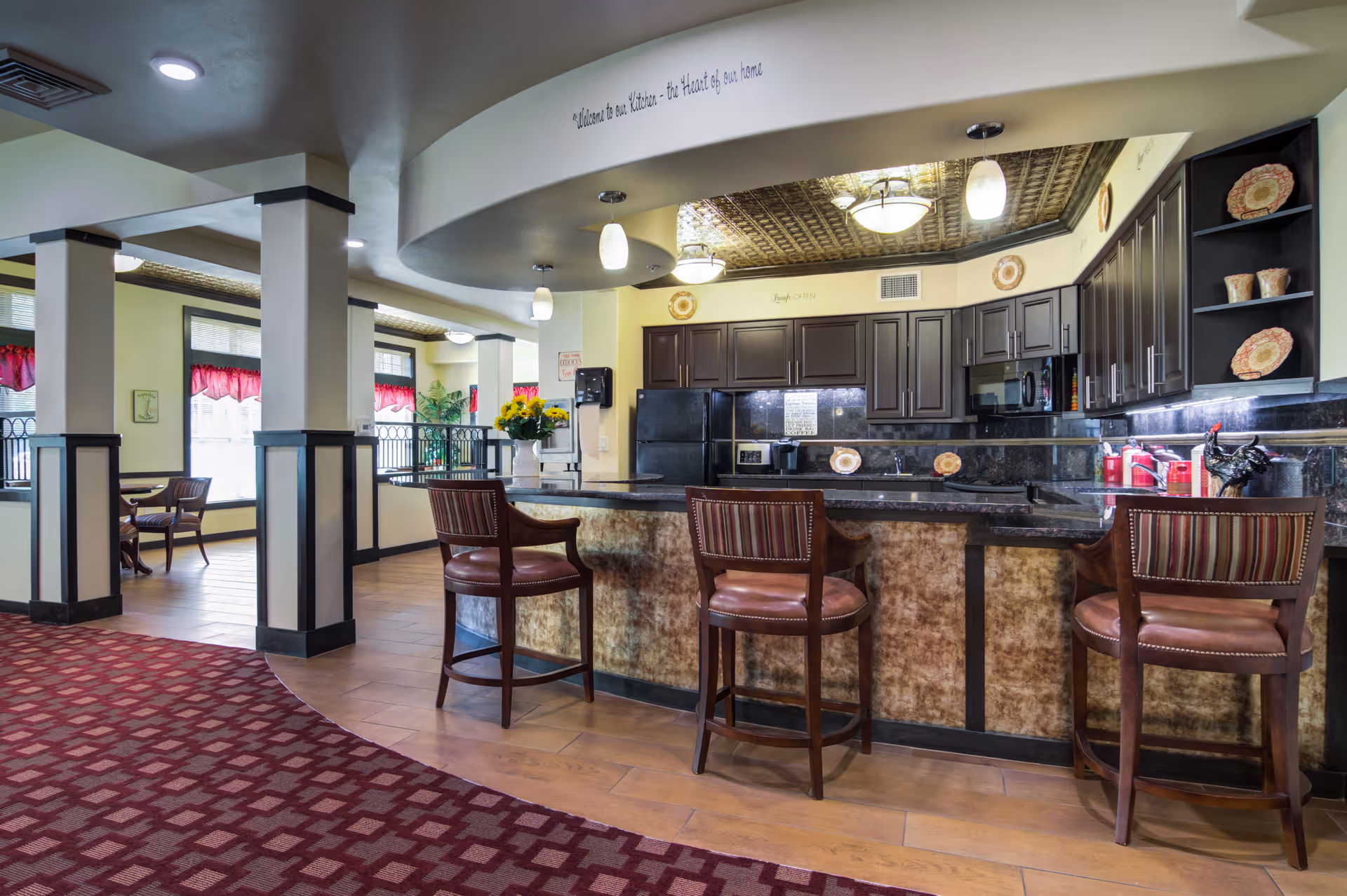 Interior view of a retirement living facility kitchen area with a curved counter and three high chairs. The kitchen has dark cabinets, a black refrigerator, and various decorative plates. The ceiling above the kitchen has a decorative pattern, and there are pendant lights hanging over the counter. Adjacent to the kitchen is a dining area with tables and chairs near windows with red valances.
