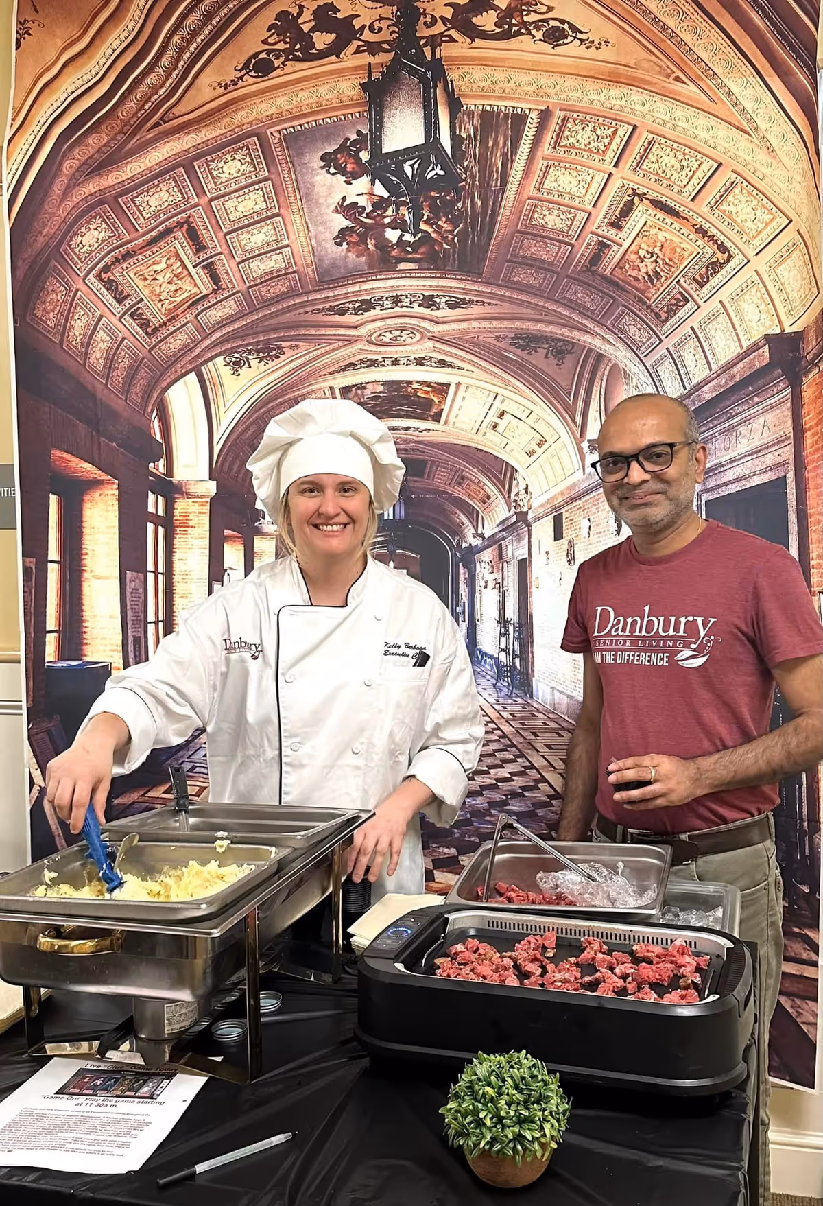 A female chef in a white chef's coat and hat stands behind a buffet table serving mashed potatoes, next to a man wearing glasses and a maroon Danbury Senior Living t-shirt. They are in front of a decorative backdrop featuring an ornate arched hallway with detailed ceiling artwork.