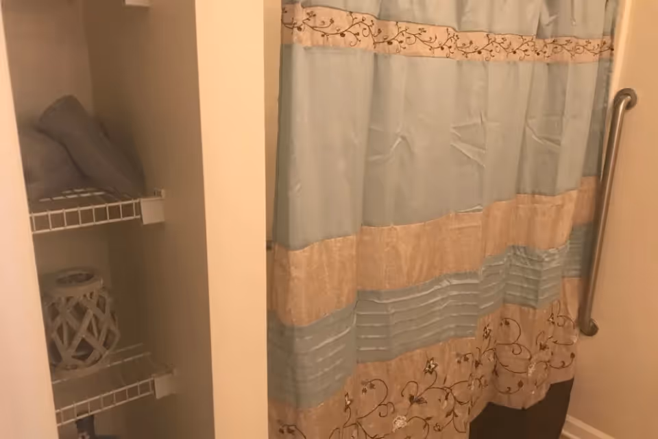 A bathroom corner showing a shower with a blue and beige floral-patterned shower curtain and a metal grab bar on the wall. To the left, there is a small open shelving unit with folded towels and a decorative item.
