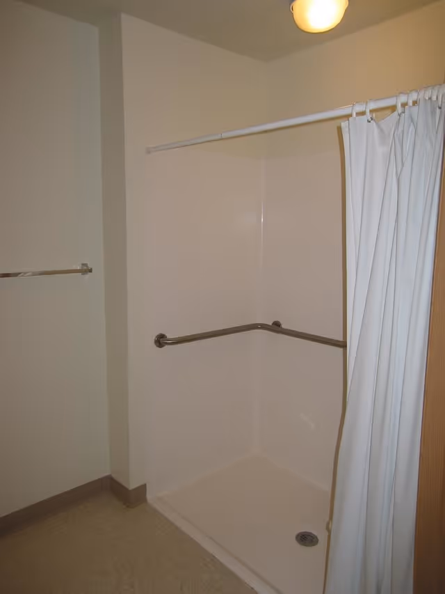 A corner shower stall with a white curtain and stainless steel grab bars in a beige bathroom.