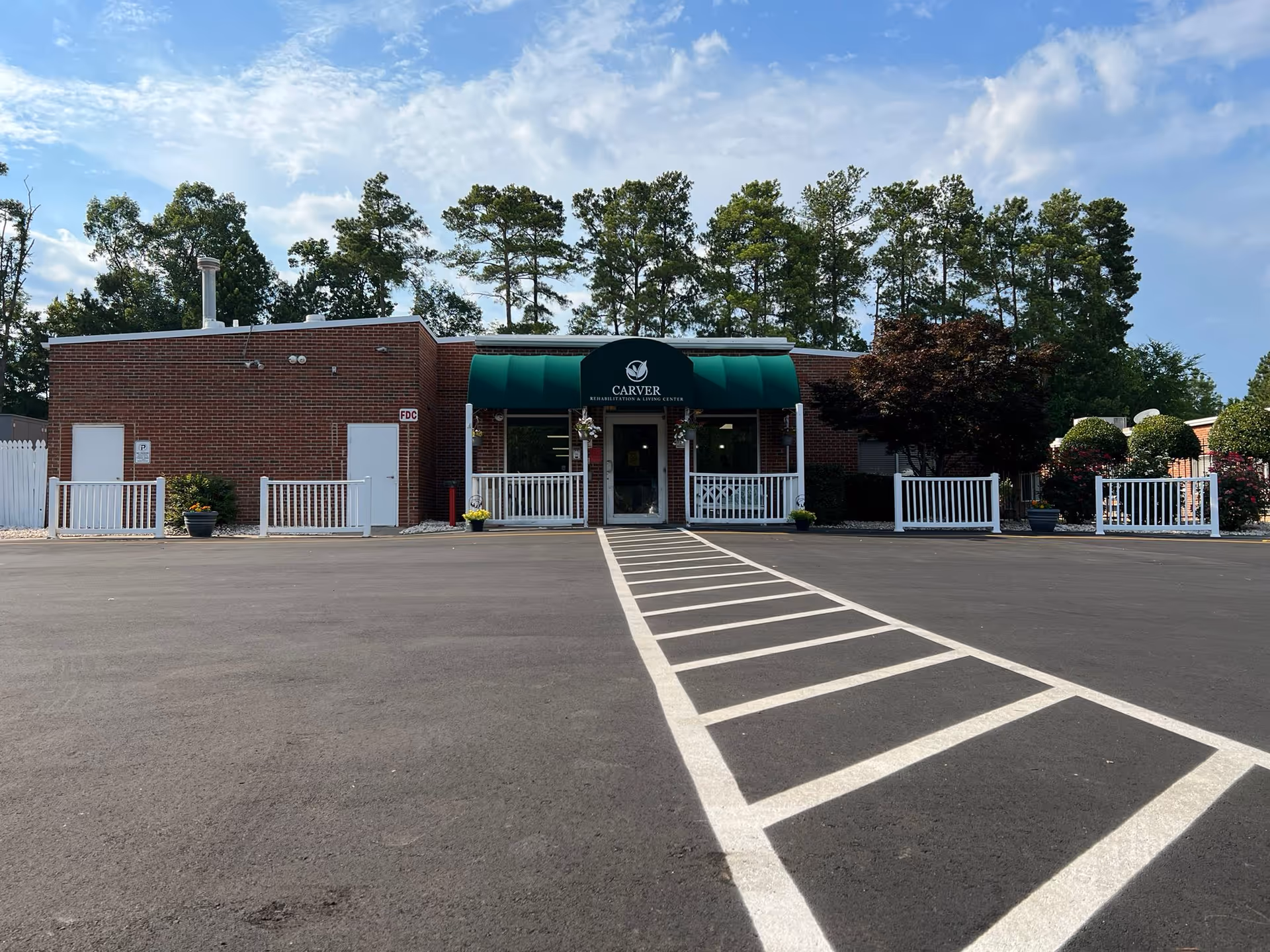 Front exterior view of Carver Rehabilitation & Living Center, a single-story brick building with a green awning above the entrance. The building is surrounded by a paved parking lot with white painted lines and some landscaping including trees and bushes.