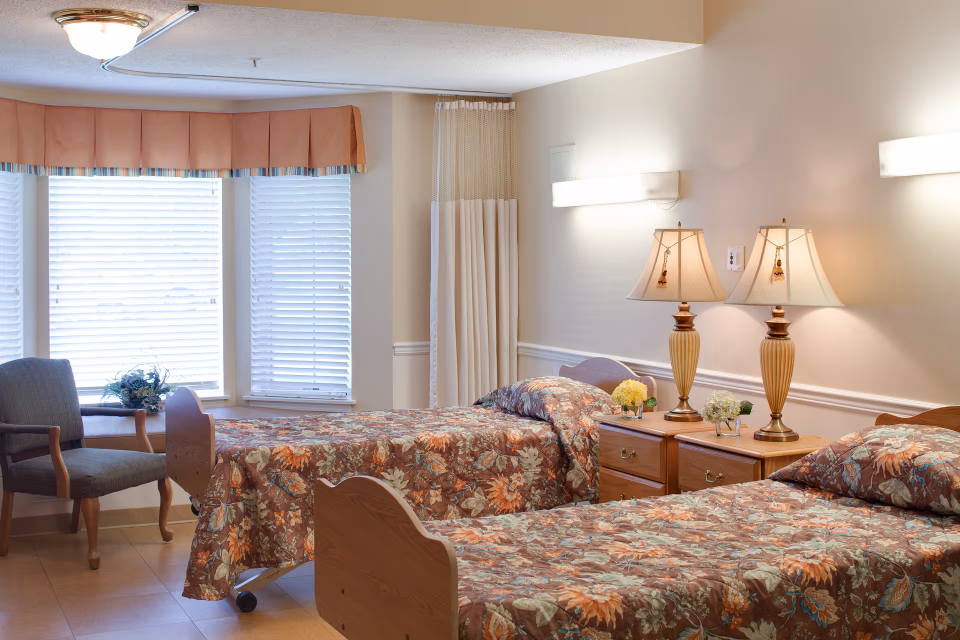 A well-lit bedroom in a senior living facility featuring two twin beds with floral bedspreads, a wooden nightstand between the beds with two matching lamps and small flower arrangements, a gray upholstered chair near a bay window with blinds, and a privacy curtain partially drawn in the corner.