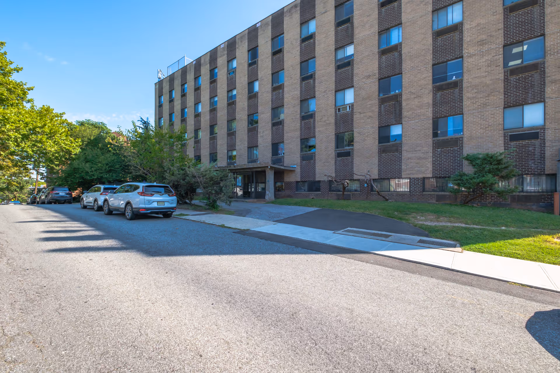 Front exterior of a multi-story brick residential building with parked cars along the street and a sidewalk leading to the entrance.