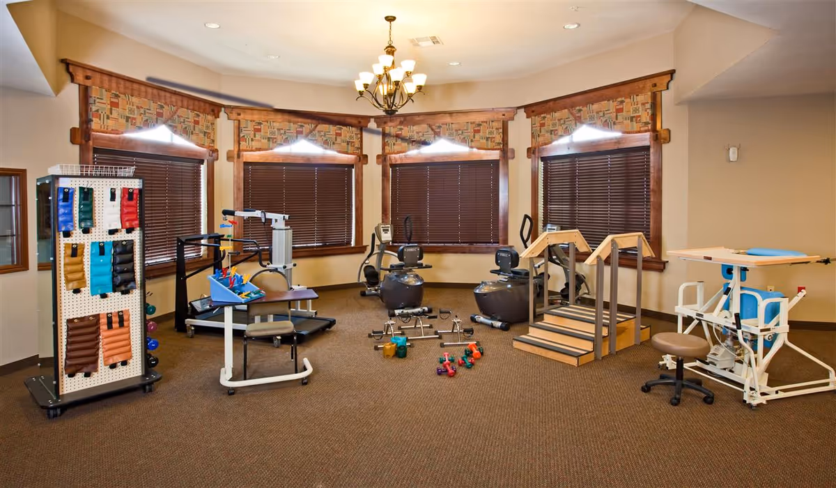 A bright interior rehabilitation room with exercise and therapy equipment, free weights, and wooden-trim windows.