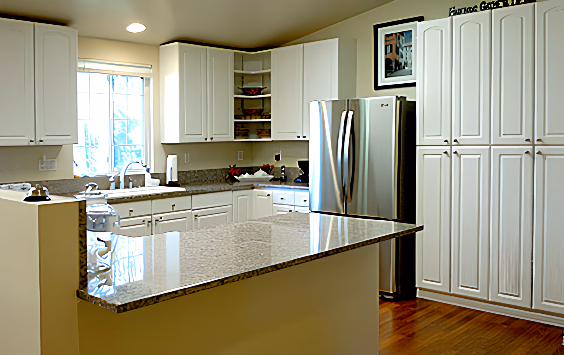 Bright modern kitchen with white cabinets, a granite island countertop, stainless steel refrigerator, and a sink under a window.