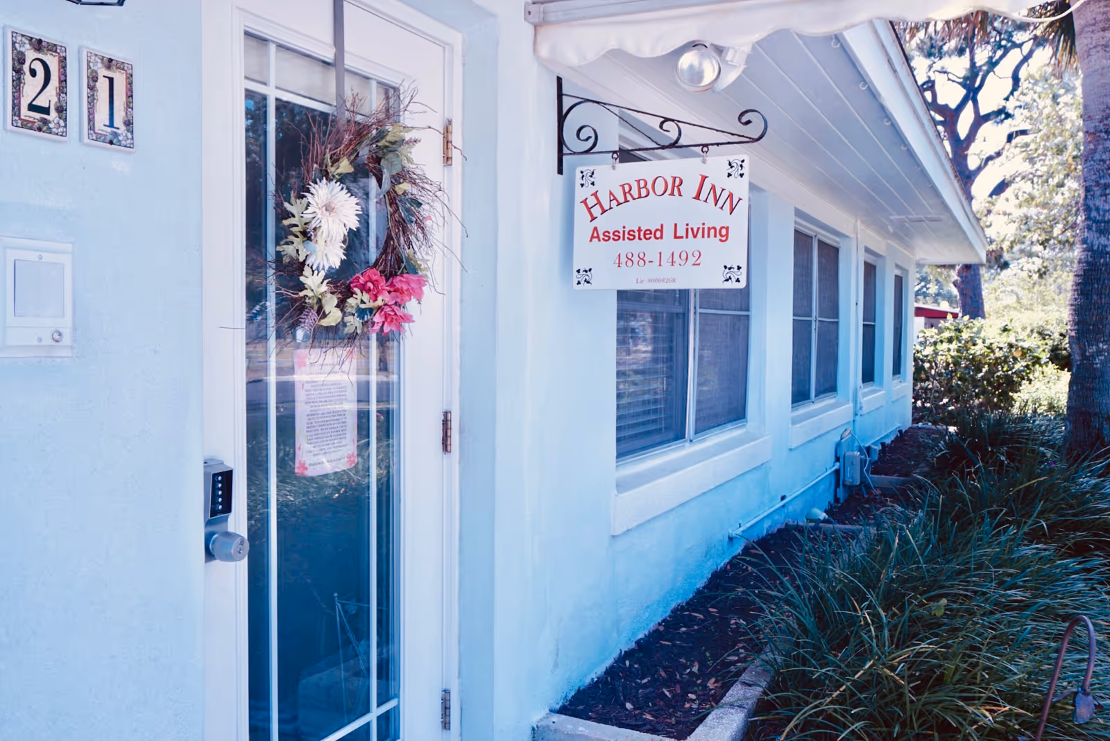 Front entrance of a pale blue assisted living building with a wreath on the glass door and a hanging sign reading 'Harbor Inn Assisted Living'.