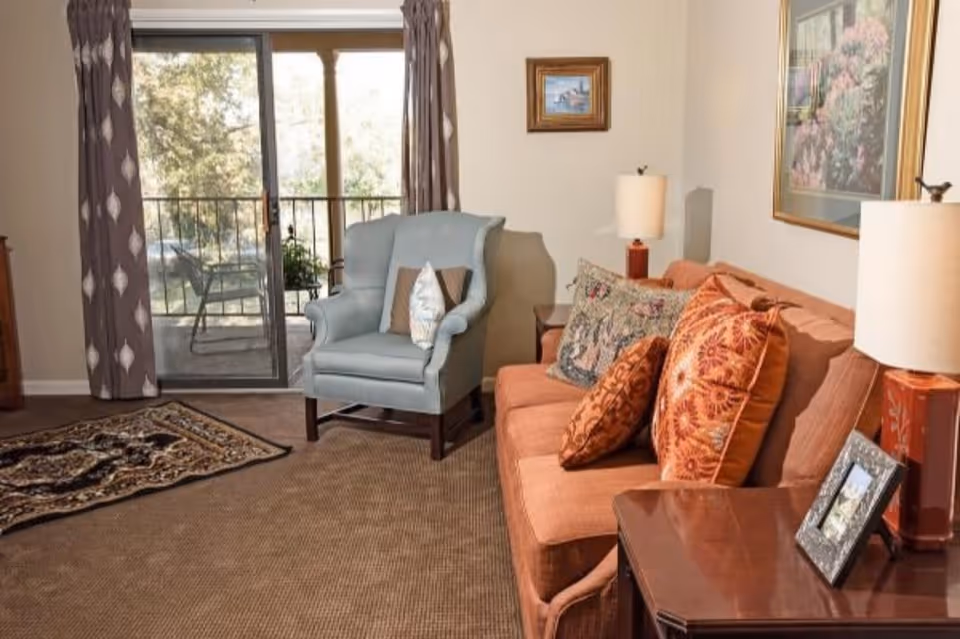 A cozy living room with a brown sofa adorned with patterned cushions, a light blue armchair with pillows, two side tables each with a lamp, framed artwork on the walls, a patterned rug on the carpeted floor, and sliding glass doors leading to an outdoor balcony with chairs and greenery visible outside.