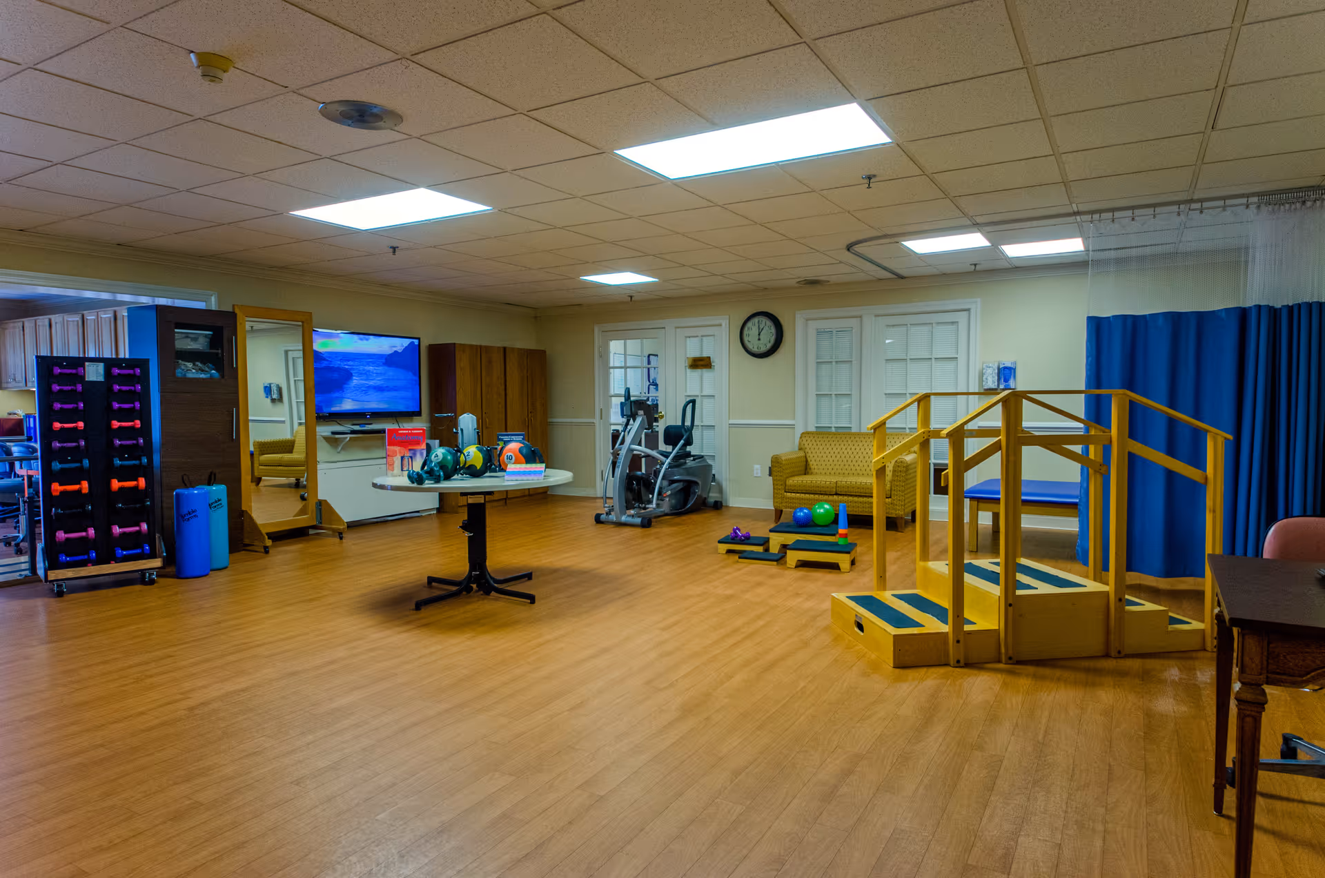 Well-lit interior therapy and fitness room with exercise equipment, weights, and a wooden rehabilitation staircase.