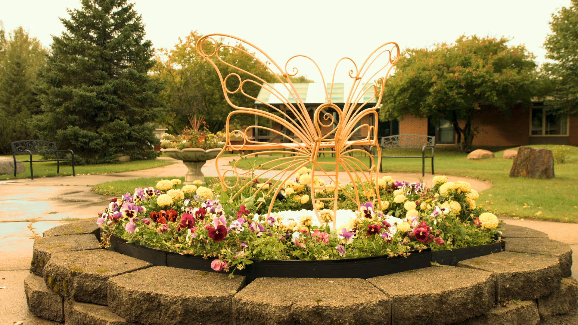 A circular raised flower bed filled with colorful pansies and a decorative metal butterfly sculpture in a landscaped courtyard with benches and trees.