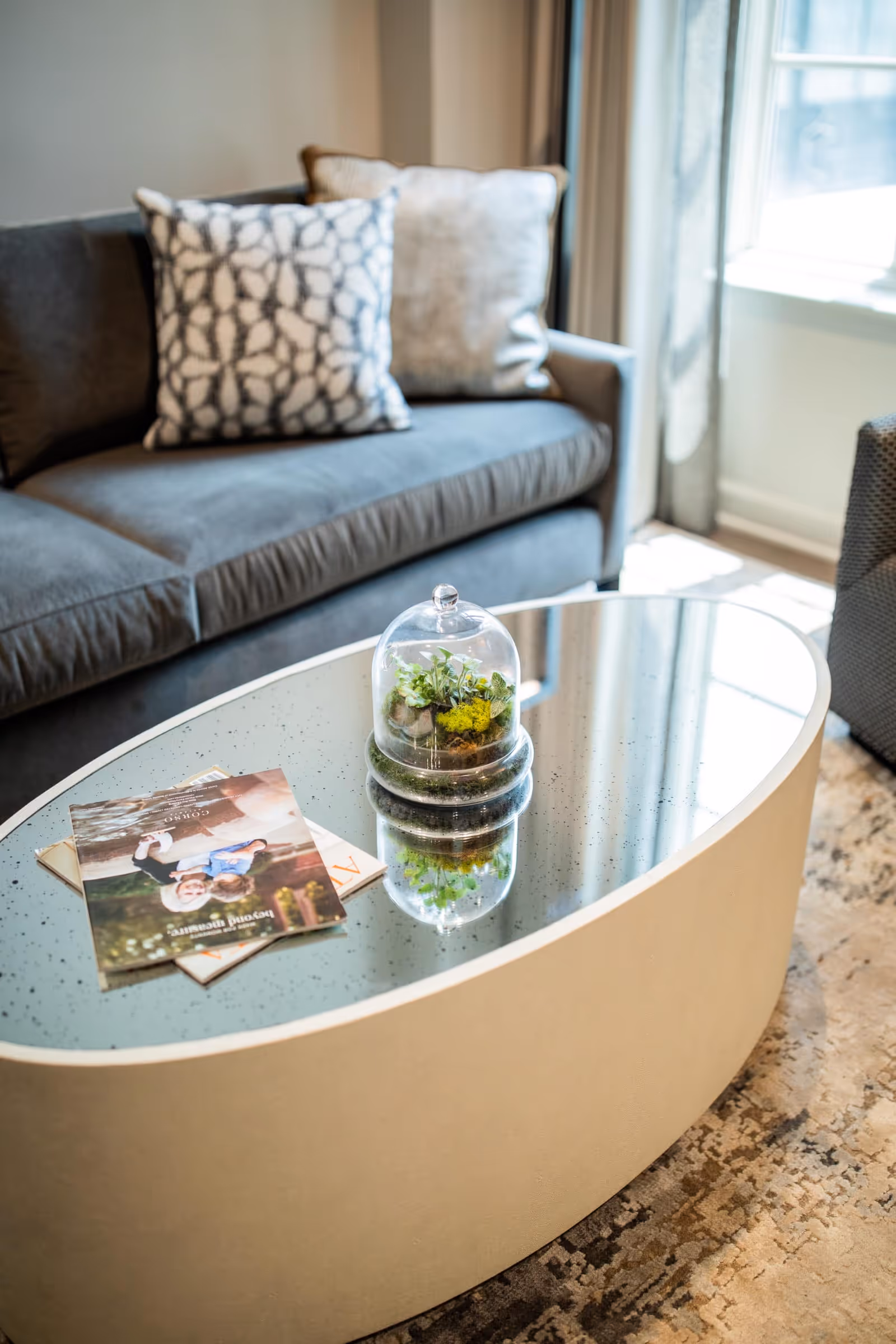 Modern living room with a gray sofa, patterned pillows, and an oval mirrored coffee table holding a glass terrarium and magazines.