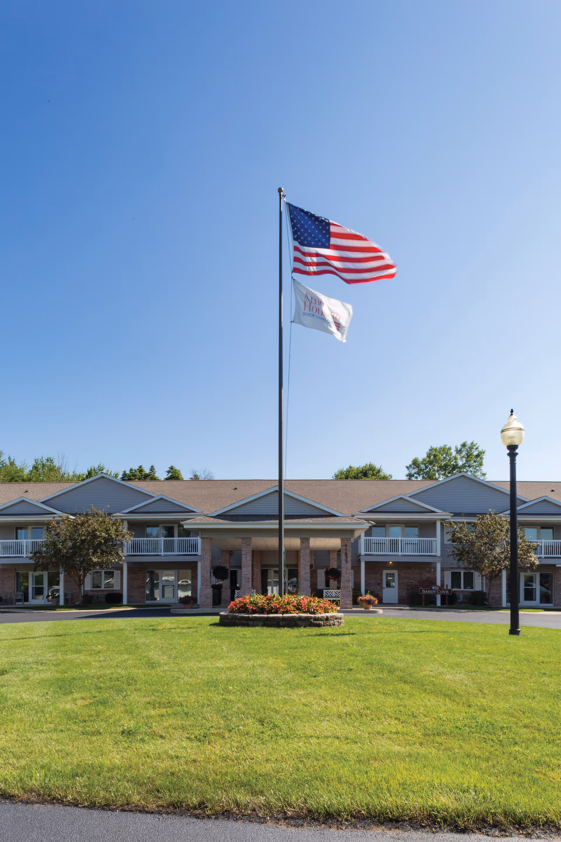 Front entrance of a two-story senior living building with a central flagpole, American flag, and a manicured lawn.