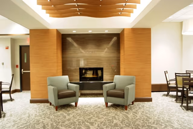 A cozy seating area in a senior living facility featuring two light blue armchairs with brown cushions positioned in front of a modern fireplace set in a wooden-paneled wall. The room has a patterned carpet and a decorative wooden ceiling feature. To the sides, there are dining tables and chairs visible, suggesting a common area or lounge.