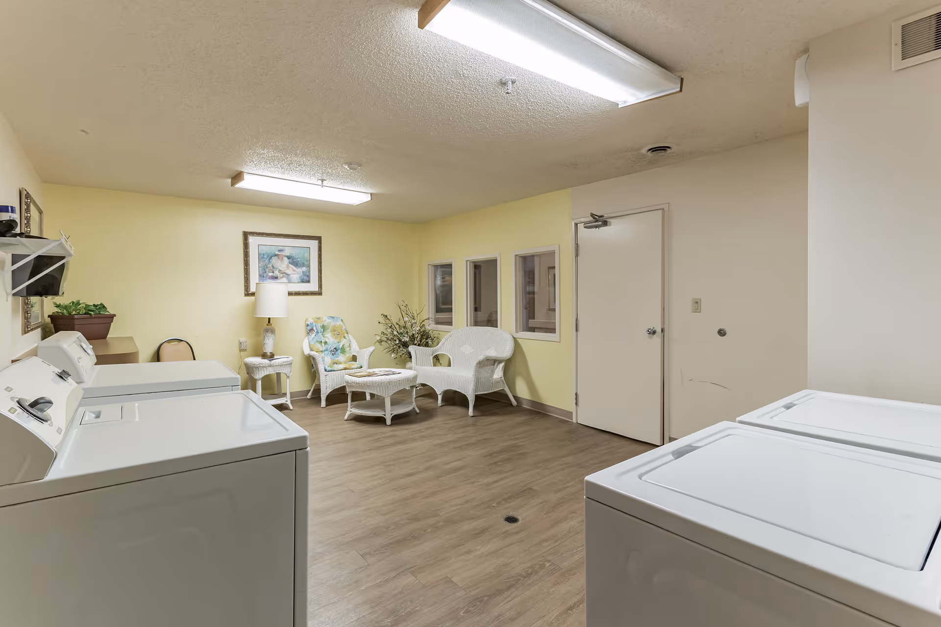 A laundry room with four white washing machines or dryers along the left and right walls. The room has light yellow walls and wood-look flooring. In the back corner, there is a small seating area with white wicker furniture including a loveseat, chair with floral cushion, coffee table, and side table with a lamp. A framed picture hangs on the wall above the seating area.
