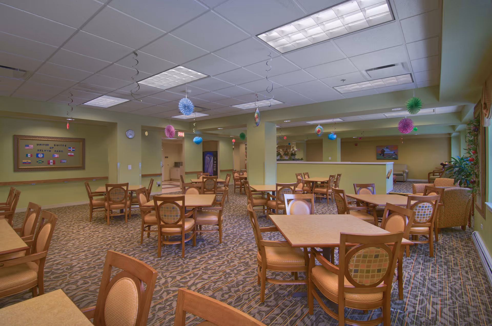 A spacious dining room with multiple wooden tables and chairs arranged neatly. The room has green walls, patterned carpet flooring, and a drop ceiling with fluorescent lights. Colorful hanging decorations are suspended from the ceiling. There is a bulletin board on one wall and a seating area with armchairs near the windows.