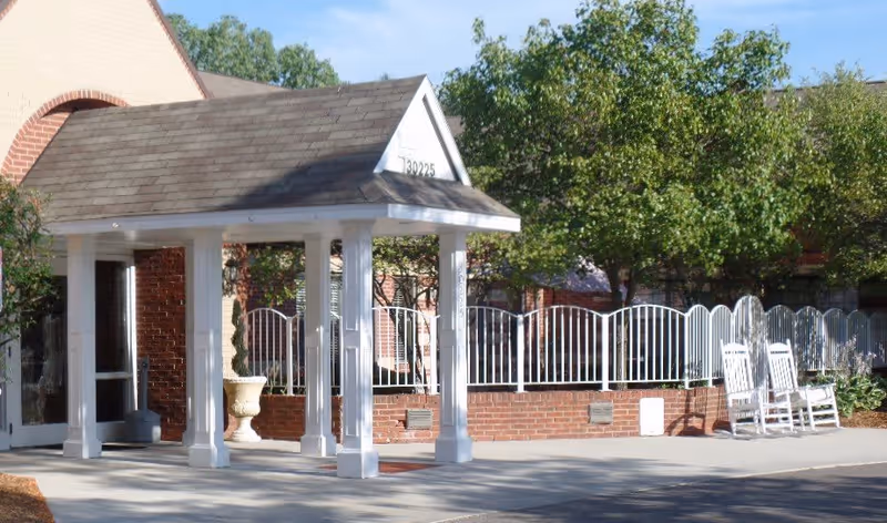 Entrance area of a building with a white covered porch supported by columns, a brick wall with white metal fencing behind it, and two white rocking chairs on the right side. Trees and greenery are visible in the background.