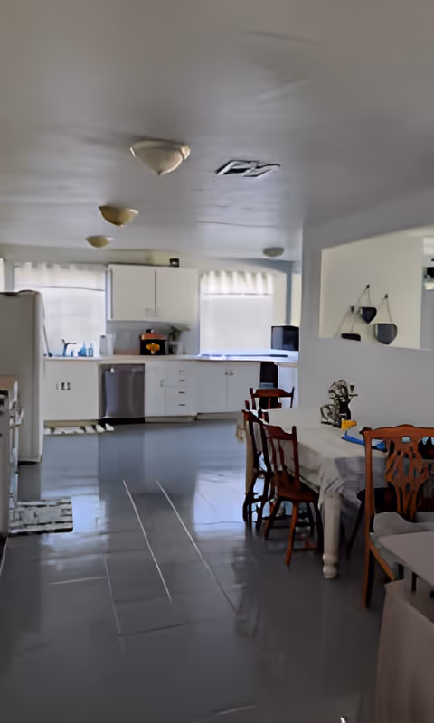Interior view of a senior living facility kitchen and dining area with white cabinets, a dishwasher, a refrigerator, a dining table covered with a tablecloth, and wooden chairs. The floor is tiled and the room is lit by ceiling lights and natural light from windows with curtains.