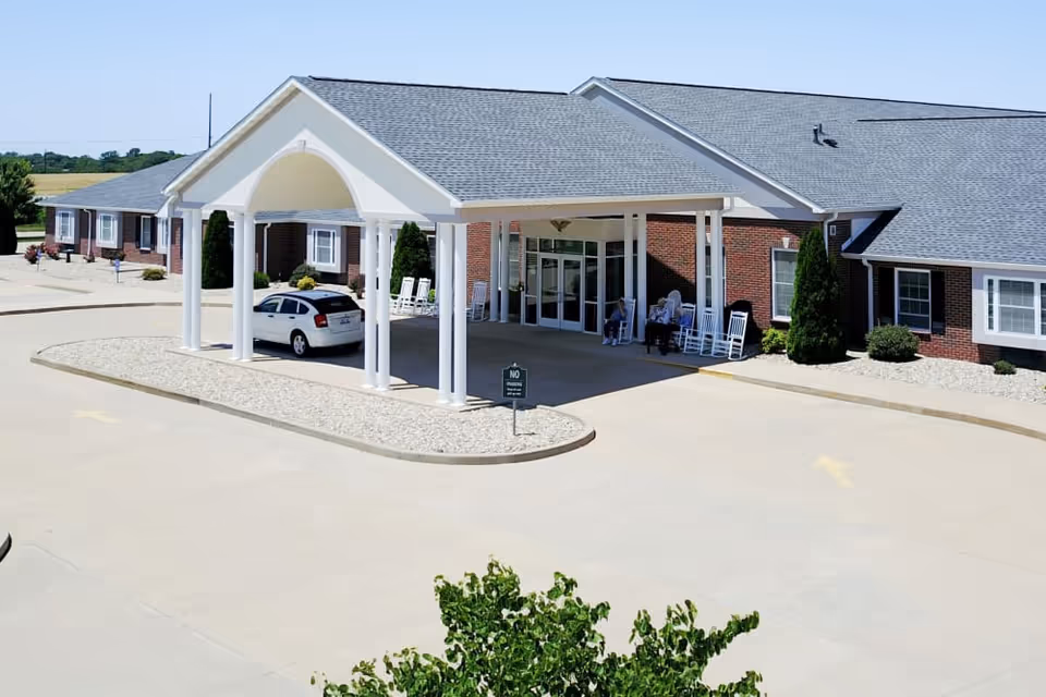 Exterior view of Garden Place Waterloo facility showing a single-story brick building with a covered entrance supported by white columns. There are rocking chairs on the porch and a few people sitting near the entrance. A white car is parked under the covered area. The surrounding area includes a paved driveway and some landscaping with bushes and rocks.